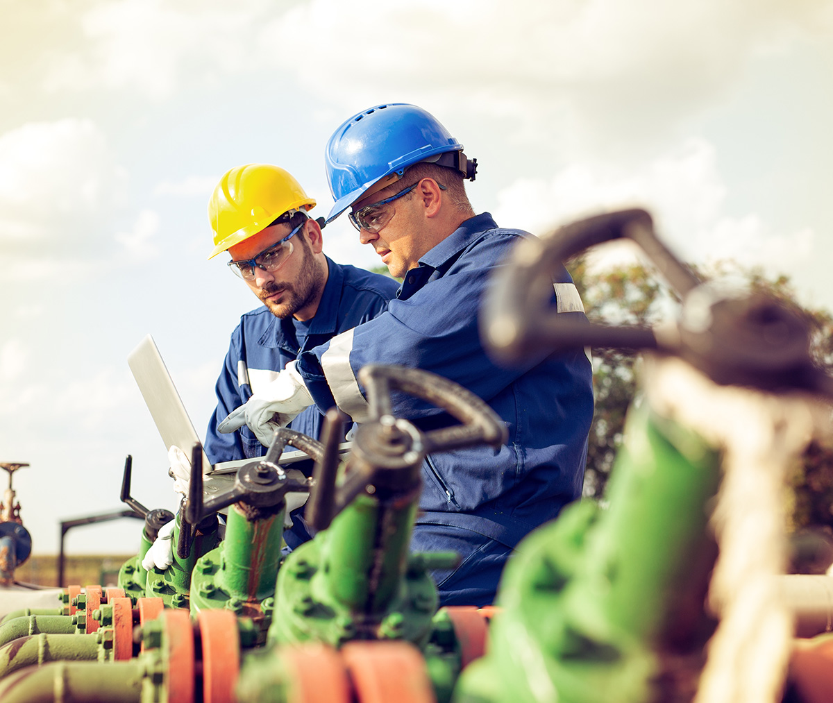 side-gas-oilrisk - VPS Learning Two workers in blue coveralls and hard hats inspect green machinery, one holding a tablet outdoors