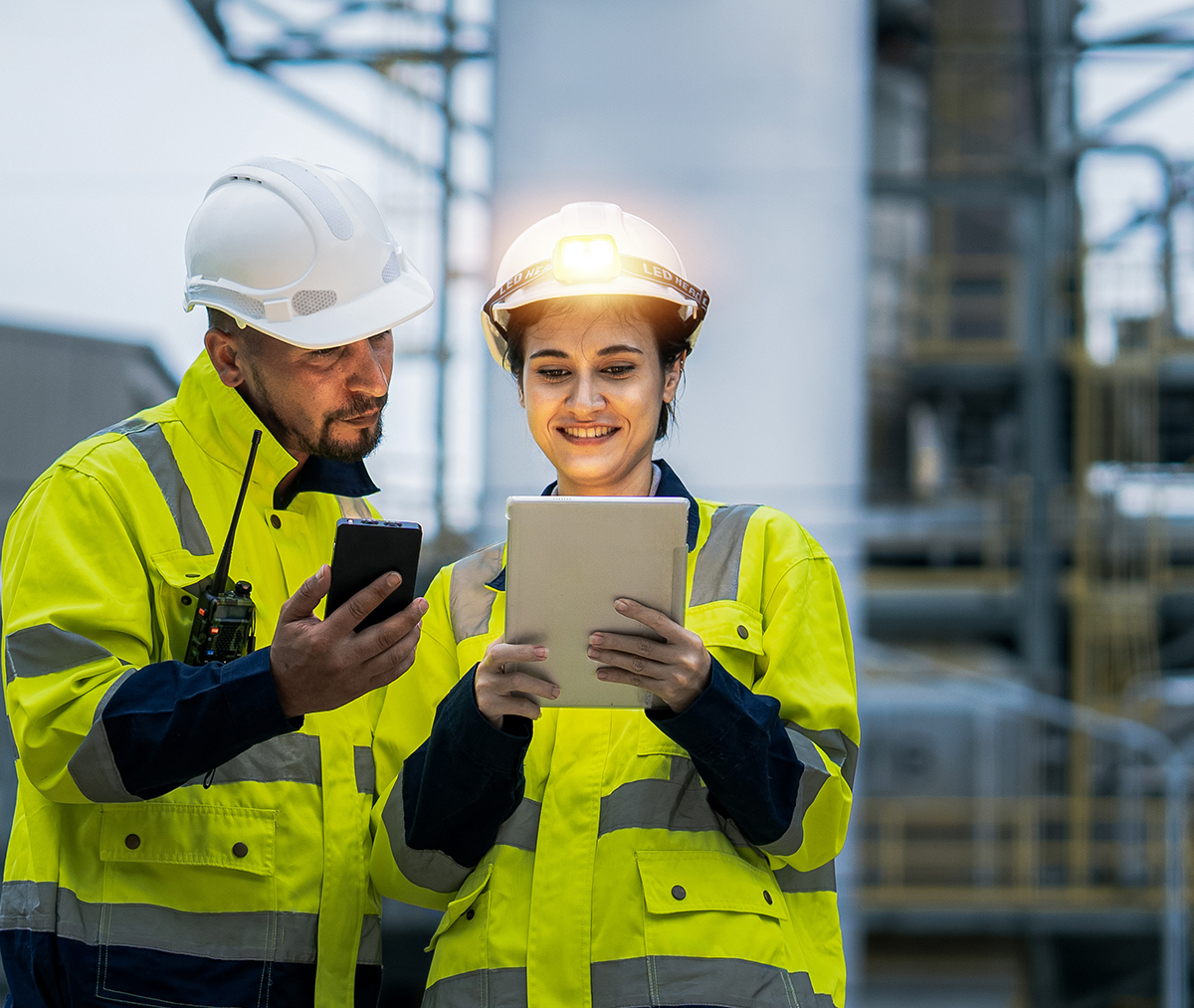 vps-energylearning - VPS Learning Two construction workers in high-visibility jackets and hard hats review a tablet and a smartphone at an industrial site.