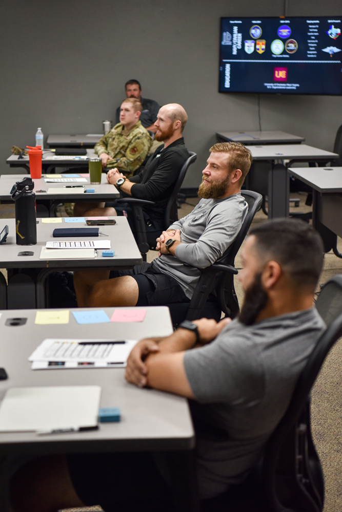The Connected Learning Environment - VPS Learning Group of men seated at desks in a training room, listening to a presentation; one in camouflage uniform, others in casual wear, with a large screen on the wall.