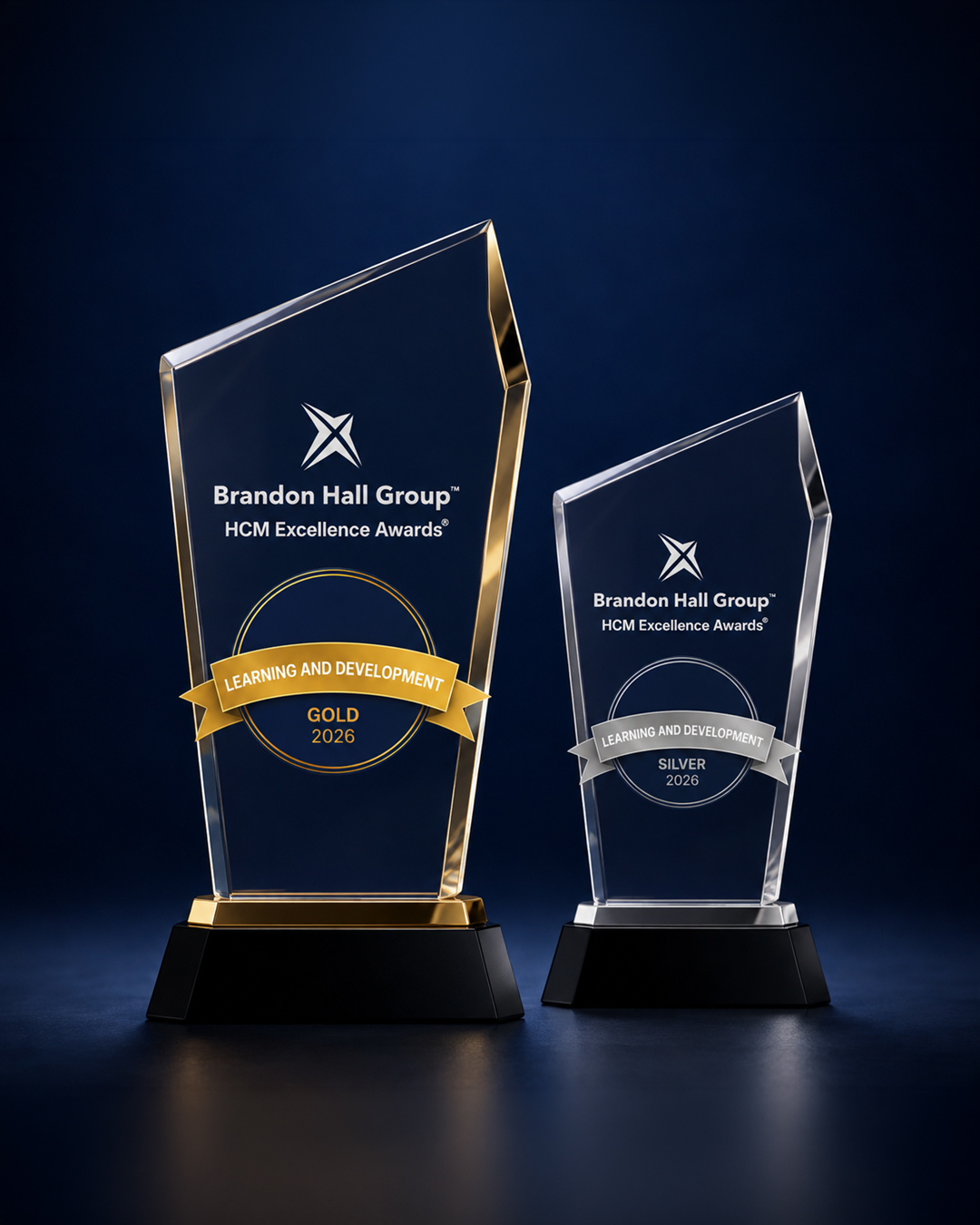 Two glass trophies for Brandon Hall Group HCM Excellence Awards (Gold 2026 and Silver 2026) on black bases, set against a dark blue backdrop.